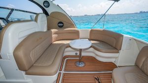 Interior seating area of a yacht with beige quilted leather benches arranged around a small round table, teak-style flooring, and bright turquoise ocean visible through the open sides.