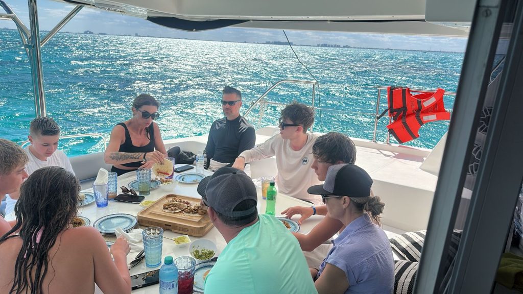 Group of adults and teenagers enjoying a meal together on a yacht, seated around a table with plates of food and drinks, turquoise ocean in the background under sunny skies.
