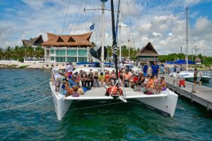 Large group of tourists seated on a white catamaran docked at a marina in Tulum, with turquoise water in the foreground and thatched-roof waterfront buildings and palm trees in the background under a partly cloudy sky.