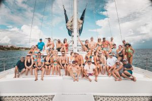 Large group of adults posing together on the deck of a catamaran at sea, wearing swimwear and sunglasses while sitting and standing around the mast with the ocean and coastline visible in the background.