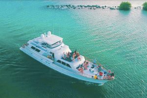 Aerial view of a large white yacht cruising through calm green-blue waters, with a group of people relaxing and socializing on the upper and front decks.