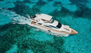 Aerial view of a luxury white motor yacht cruising over clear turquoise Caribbean water, with coral reef patterns visible beneath the surface and a small wake trailing behind the vessel.