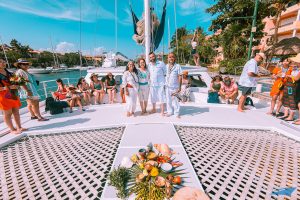 Group of guests dressed in light, festive attire standing and sitting on a catamaran docked at a marina in Tulum