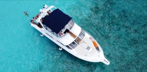 Top-down aerial view of a white yacht anchored in clear turquoise water, with several people relaxing on deck and one person swimming nearby.