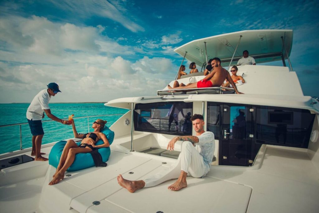 Group of guests relaxing on a luxury white yacht in turquoise Caribbean waters, with a crew member serving drinks while others lounge on the upper deck under a bright blue sky.