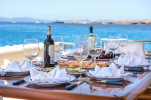 A wooden table set for an outdoor meal on a yacht, with wine bottles, empty wine glasses, folded white napkins, sliced bread, fruit, and appetizers, overlooking bright blue ocean water in the background.
